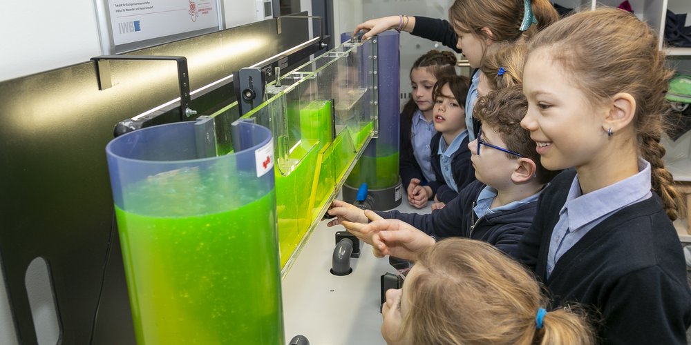 Staunende Kinder blicken auf einen großen Glasbehälter mit grüner Flüssigkeit.