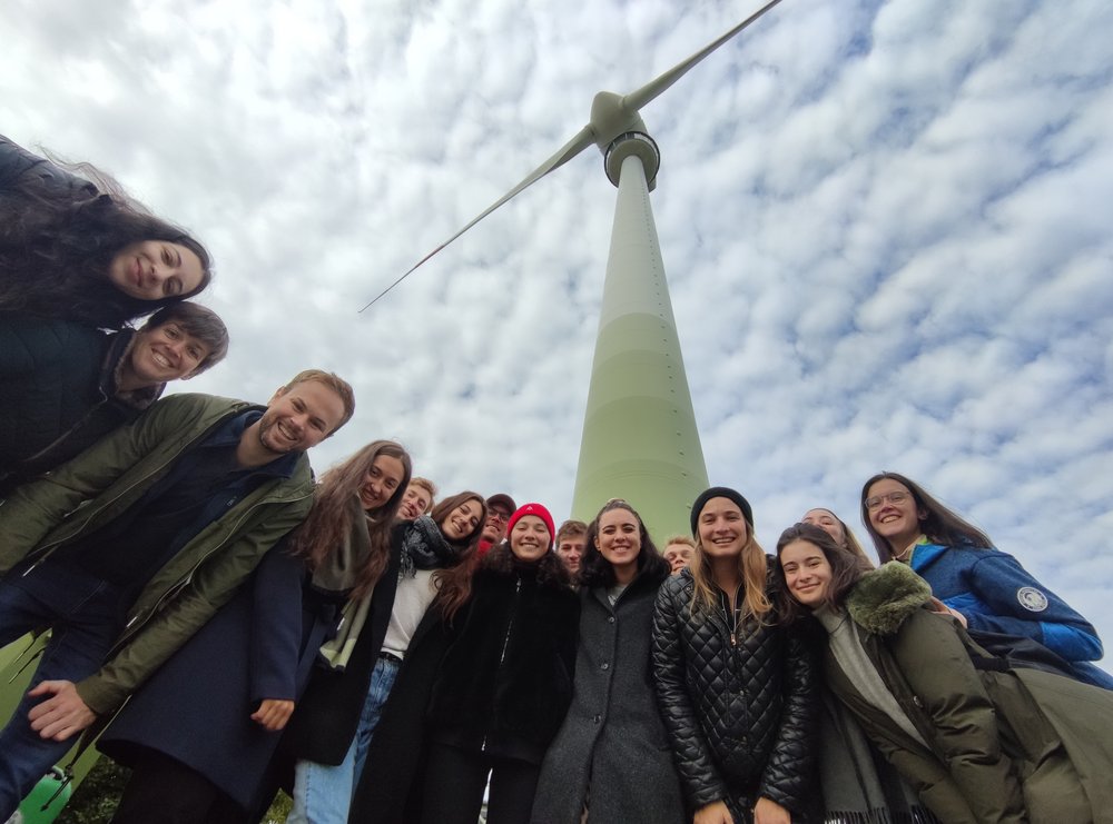 Gruppe von Menschen von unten fotografiert, dahinter ragt ein Windrad auf; im Rahmen einer Exkursion des MSc Environmental Technology & International Affairs (ETIA) zu den Windrädern