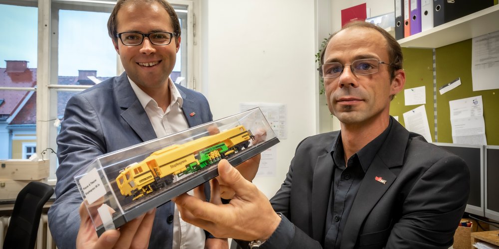 Zwei TU Graz-Forschende mit einem Modell einer Stopfmaschine in Händen