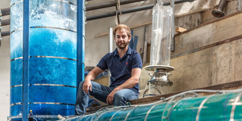 Ein junger Mann mit Bart in Jeans und blauem T-Shirt sitzt im Wasserbaulabor und ist umgeben von Versuchanordnungen in Form von wassergefüllten Plexiglas-Röhren.