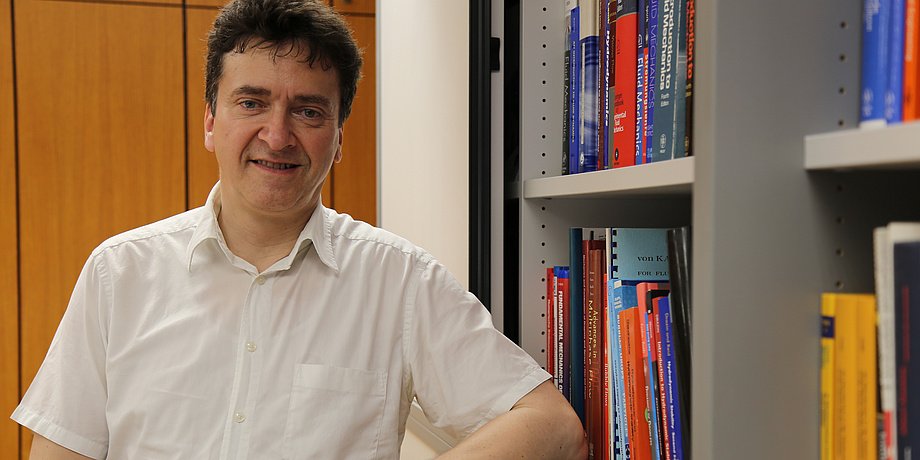 Brown haired man with short-sleeved white shirt standing in front of a wooden cupboard with his elbow on a bookshelf.