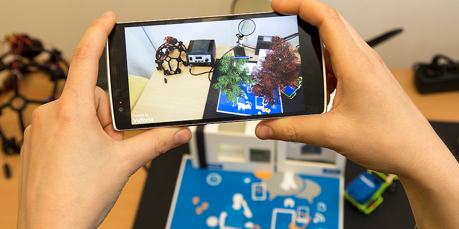 Ein Smart-Phone wird von zwei Händen gehalten. Am Bildschirm zu sehen ist die Visualisierung eines Versuchaufbaus am Know-Center.