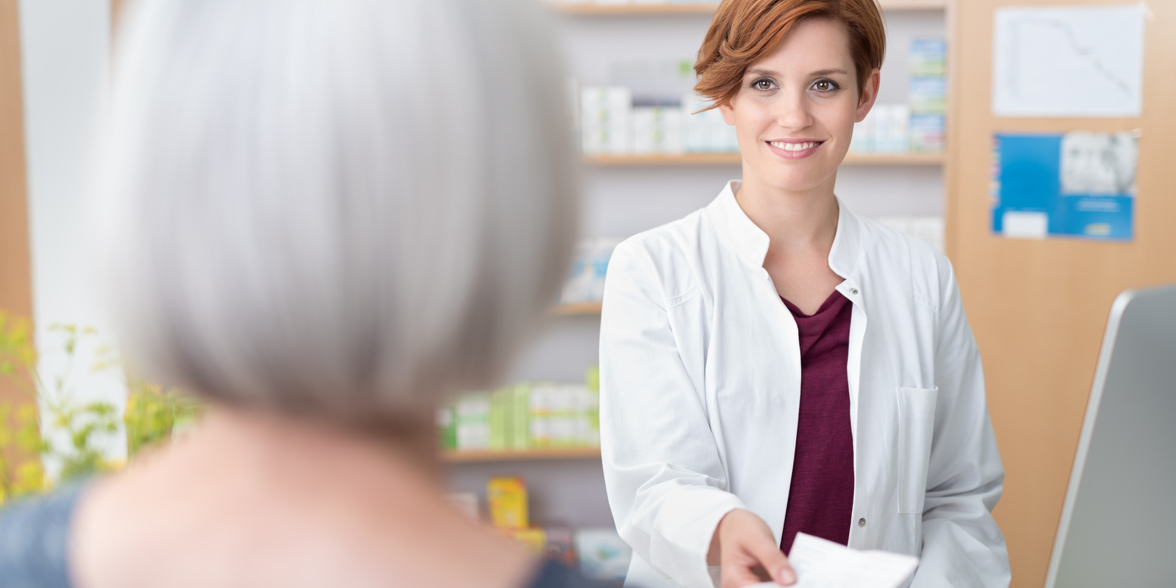 A grey haired lady gives a prescription to a young pharmacist.