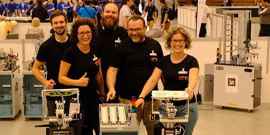 Three men and two women wearing black Shirts with TU Graz and GRIPS Logos showing proudly robots of the der Logistics League.